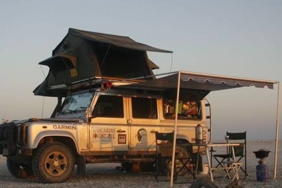 A white Land Rover Defender set up with a rooftop tent and side awning at a bush camp.