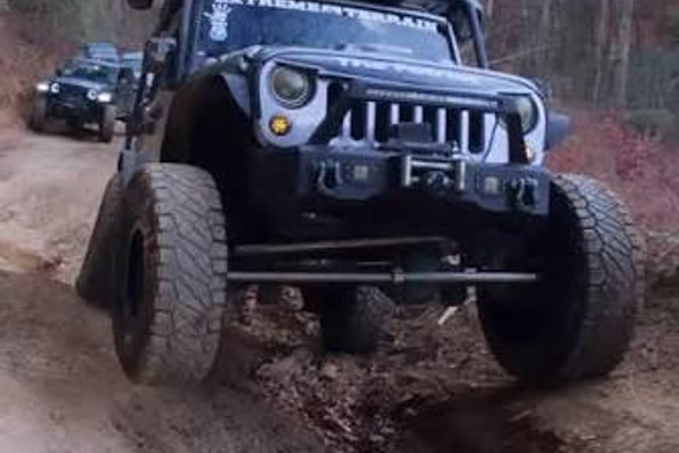 A black Jeep Wrangler climbing a rutted forest trail, a second Jeep behind it.