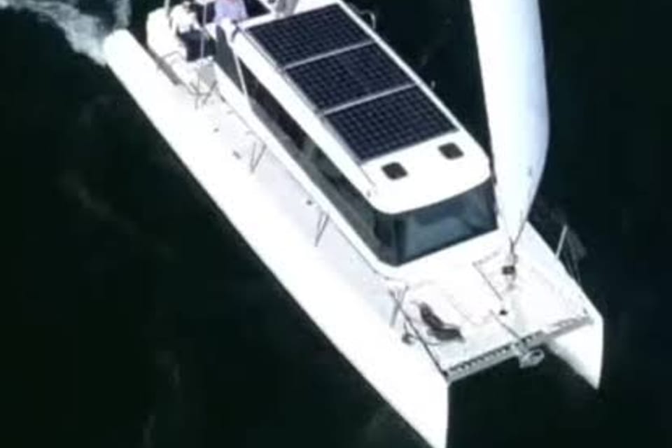 A compact cruising sailing catamaran seen from above, deck solar panels fitted, under way on dark water.