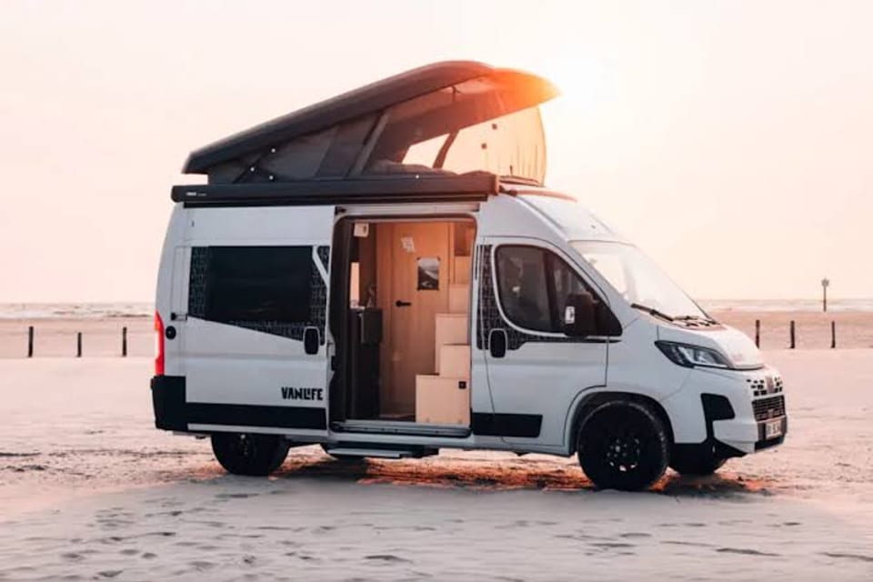 A white pop-top camper van parked on a beach at sunset, side door open.