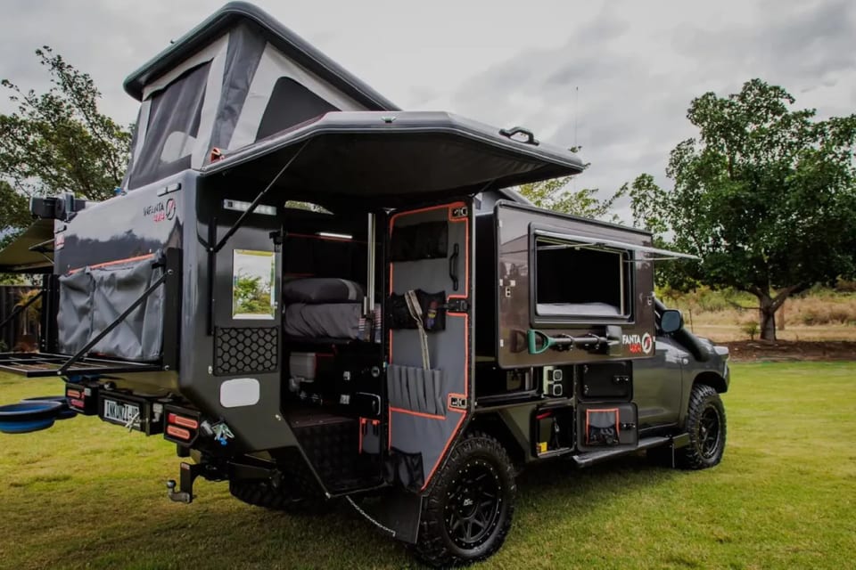 A black 4x4 pick-up carrying a slide-in pop-top camper, opened up at a grassy campsite.
