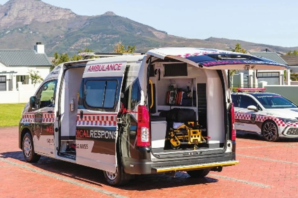 An emergency-response ambulance van with its rear open showing a stretcher, a response car behind, mountains in the background.